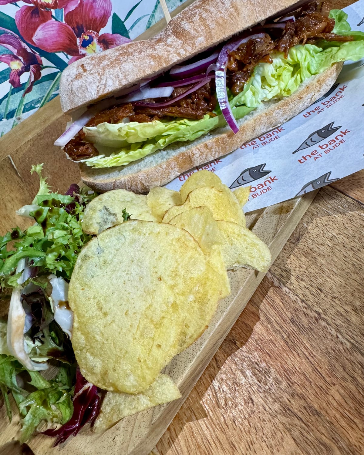 A BBQ pulled pork ciabatta with red onion and salad leaves, served with crisps and salad on a board at The Bank at Bude