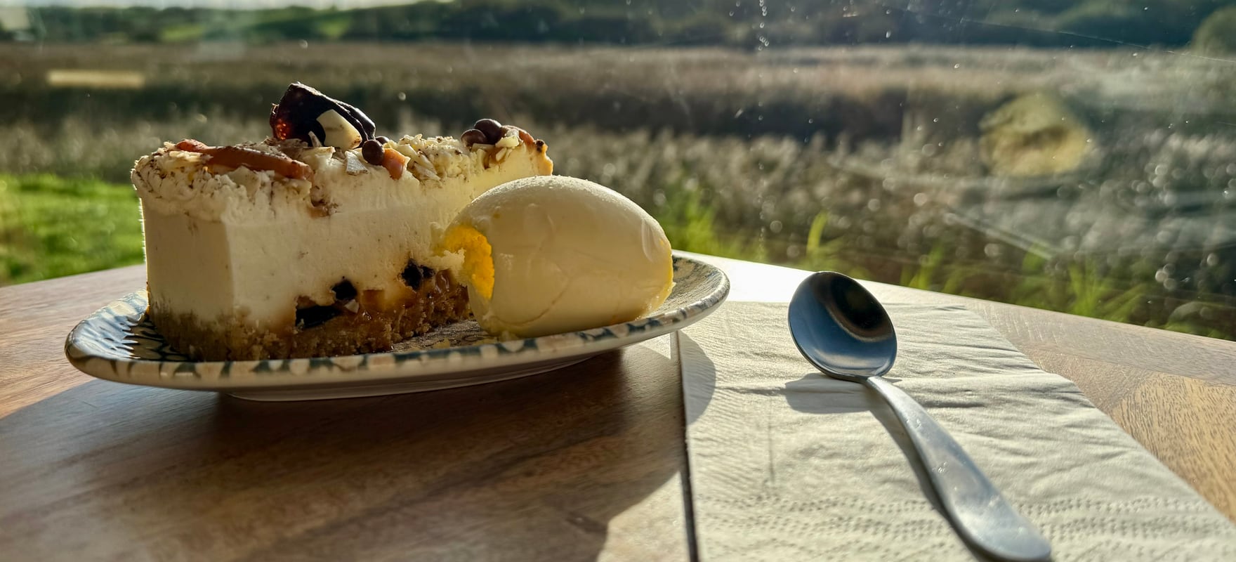 A slice of cheesecake with ice cream on the table looking out over the River Neet marsh at golden hour