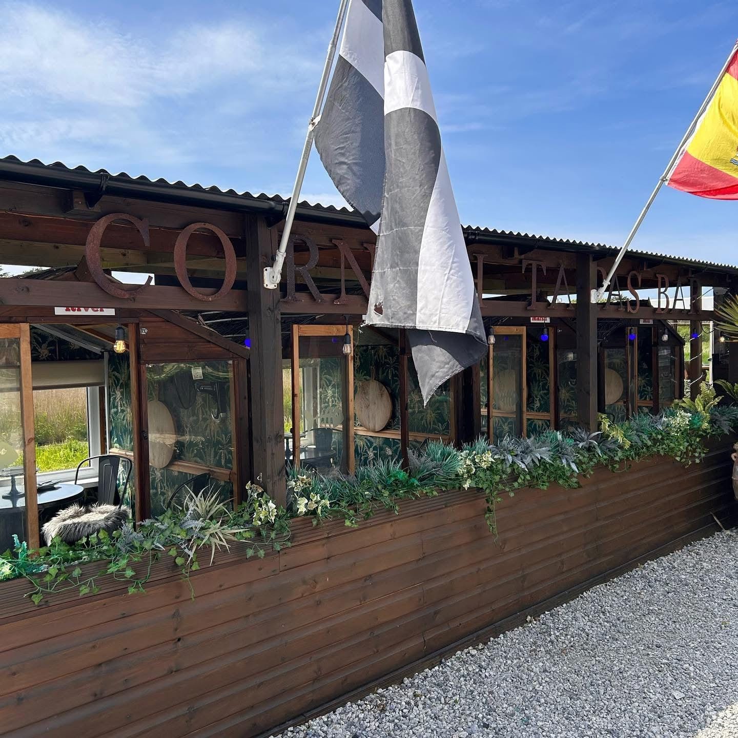 The row of riverside dining pods at The Bank at Bude beneath the ‘Cornish Tapas Bar’ sign, flying the Cornish and Spanish flags beside the River Neet marsh