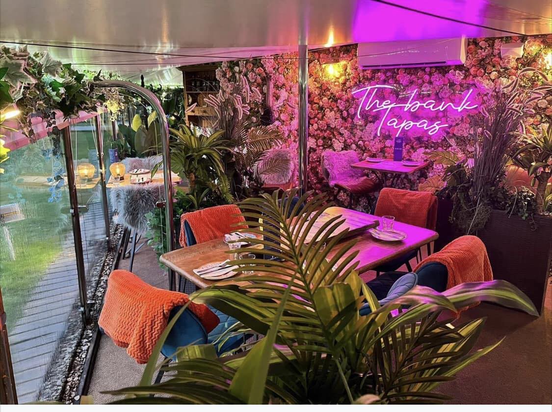 The Bank at Bude interior — pink neon ‘The Bank Tapas’ sign above set tables and a floral wall
