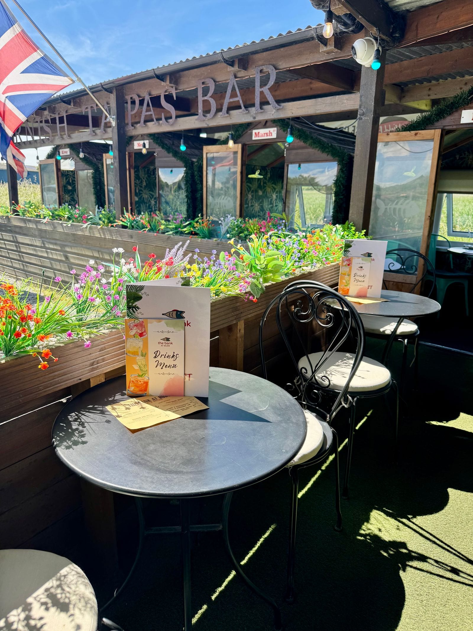 Bistro tables and ironwork chairs on the sunny outdoor terrace at The Bank at Bude beneath the Tapas Bar sign