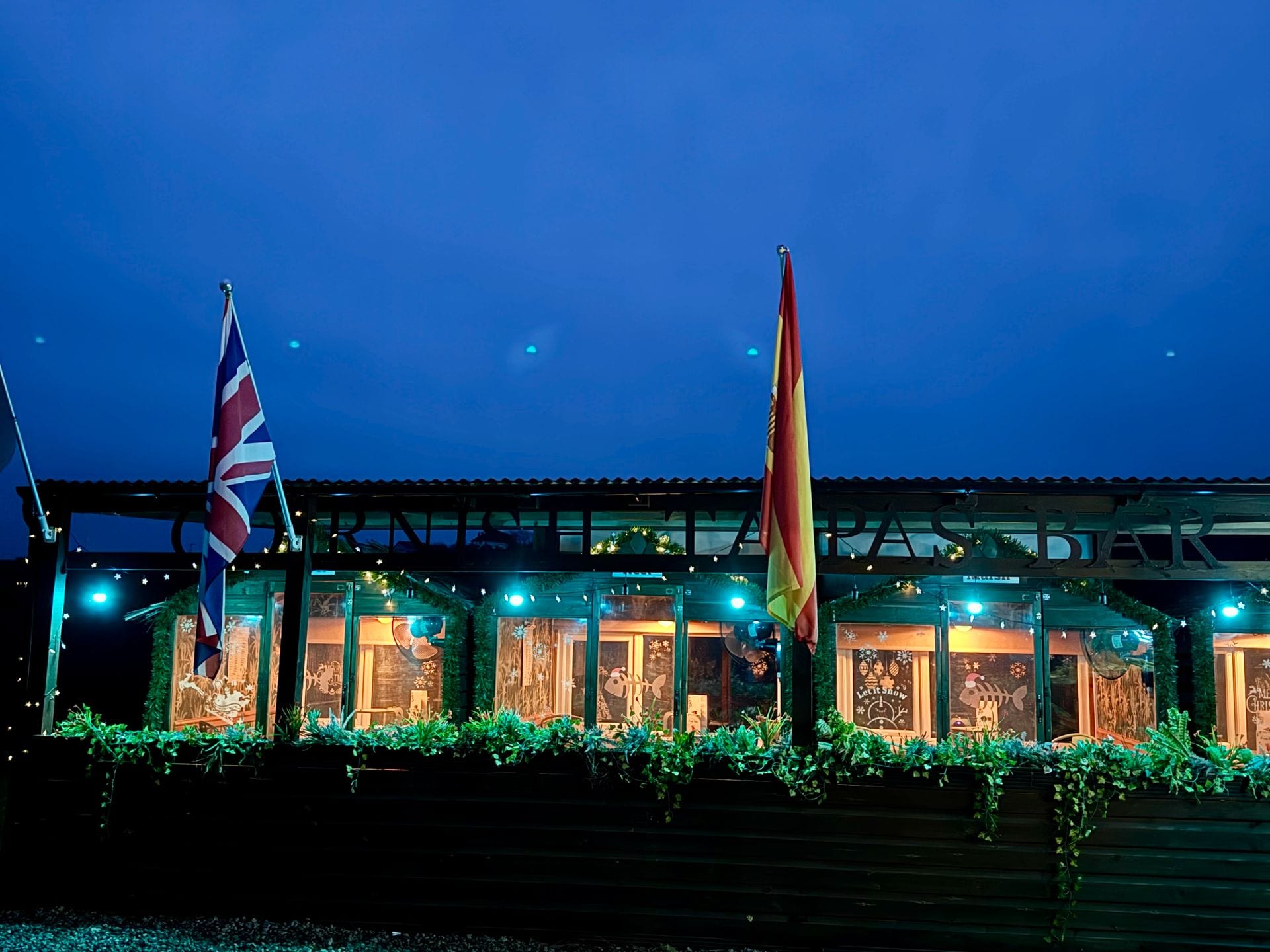 The row of riverside dining pods at The Bank at Bude lit at blue hour beneath the Cornish Tapas Bar sign