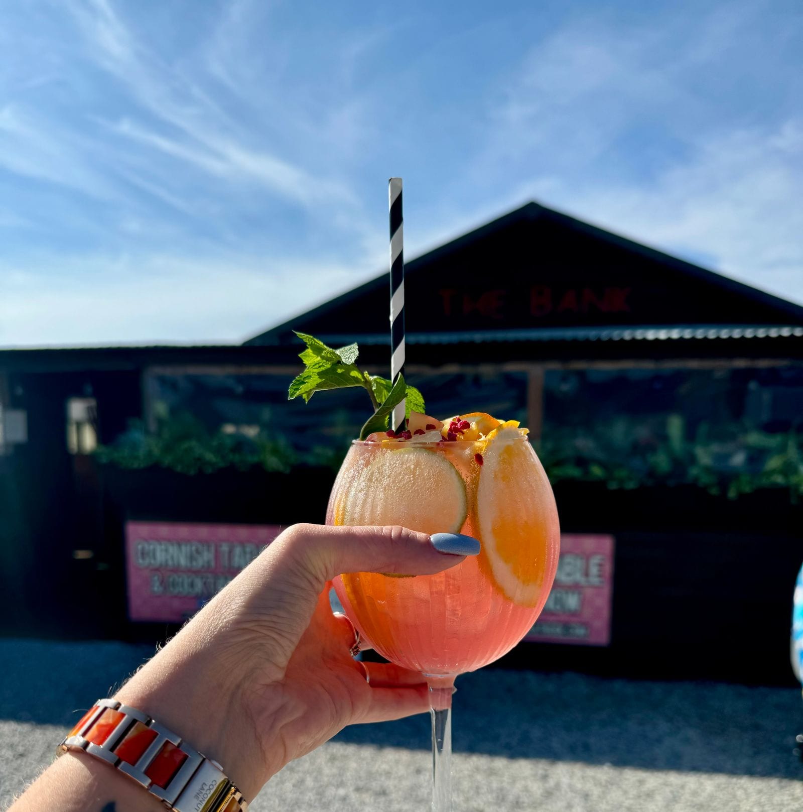 A signature fruit cocktail served in a balloon glass in front of The Bank at Bude’s lit sign