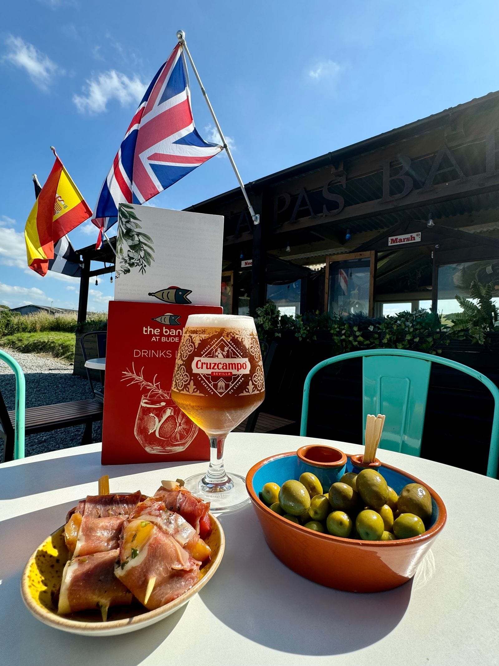 Spanish tapas at The Bank at Bude — marinated olives, jamón and a glass of Cruzcampo in the sun beneath the Tapas Bar sign, with the Cornish, Spanish and Union flags