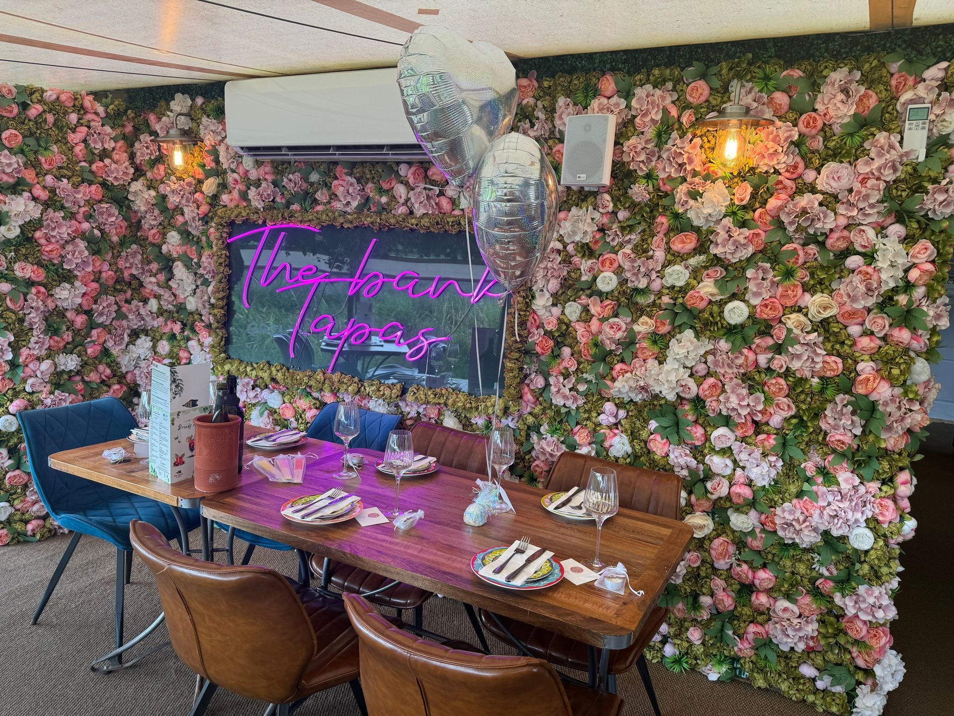 A long table set for a private celebration at The Bank at Bude against the pink-neon ‘The Bank Tapas’ floral wall, with balloons and Champagne